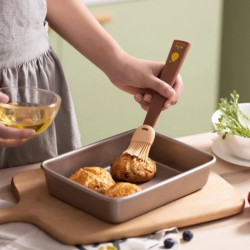 Person brushing food in a baking dish with a brush, holding a bowl of oil.