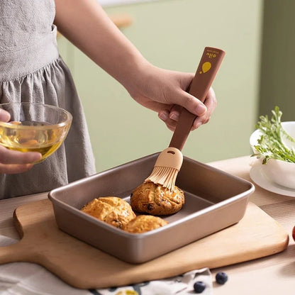 Person brushing food in a baking dish with a brush, holding a bowl of oil.