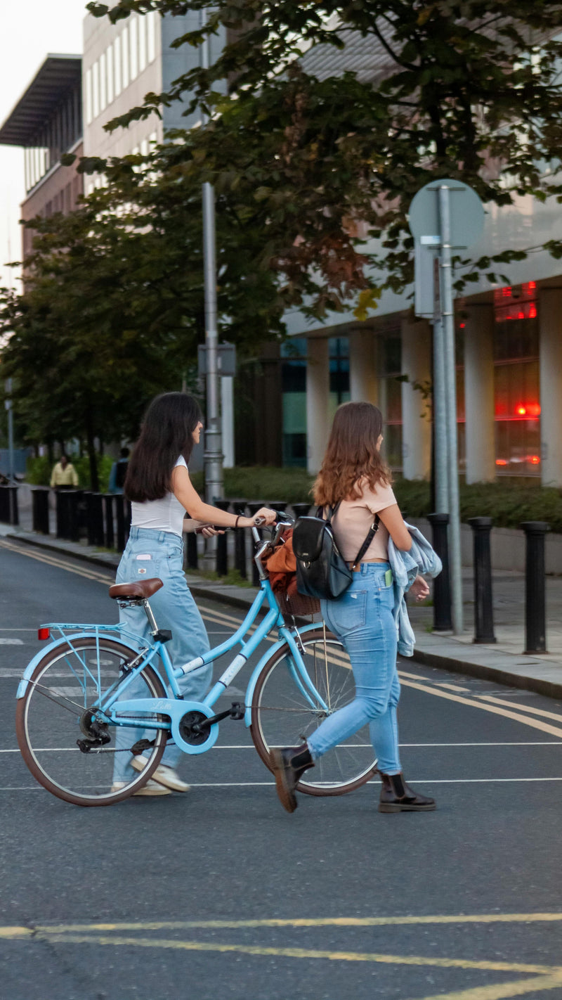 Two women walking with a light blue bicycle on a city street.